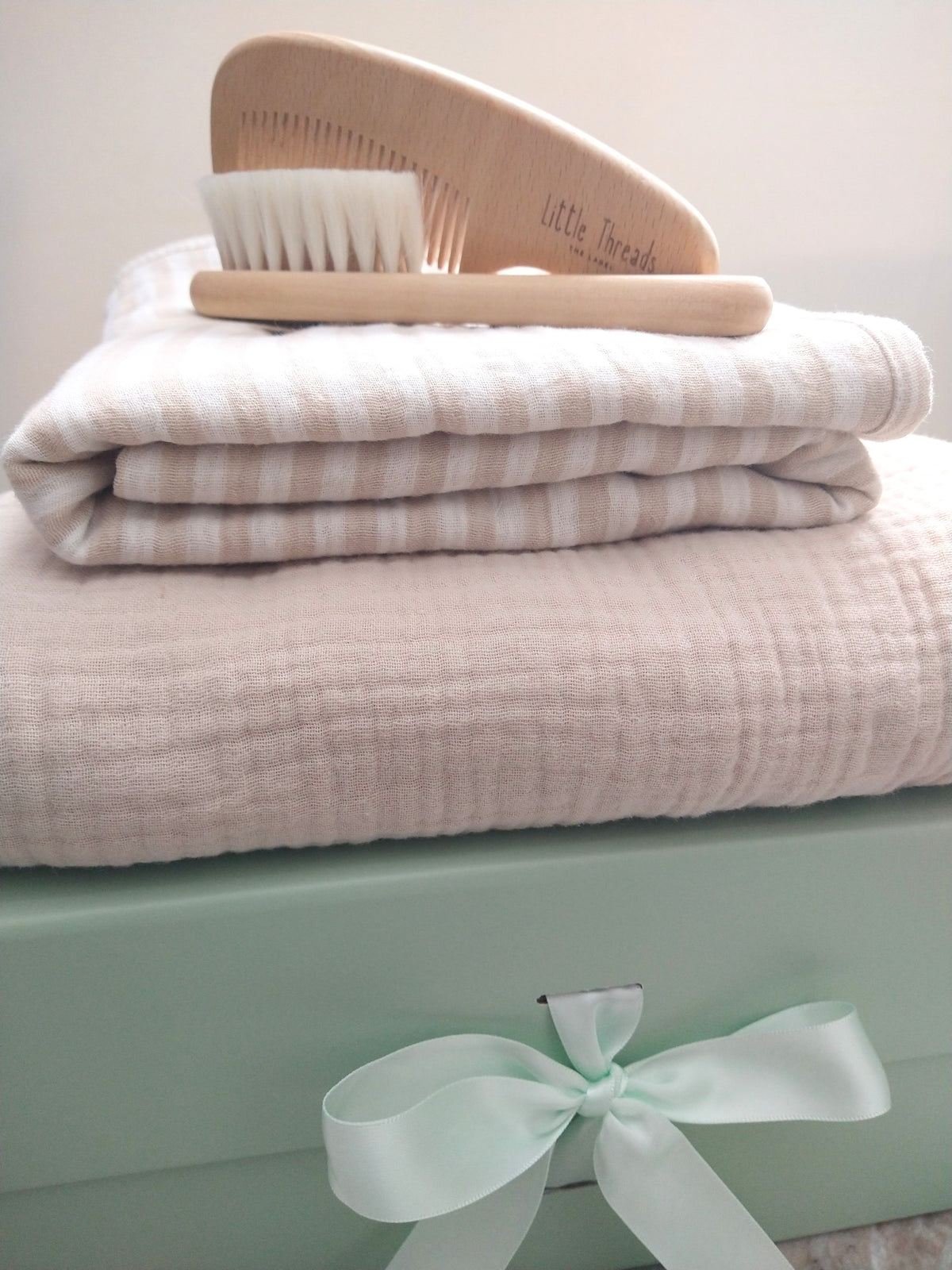 Petite Evelina Light Beige hooded towel, Beige Stripes swaddle and Little Threads wooden brush and comb displayed on sage green gift box with coordinating ribbon.