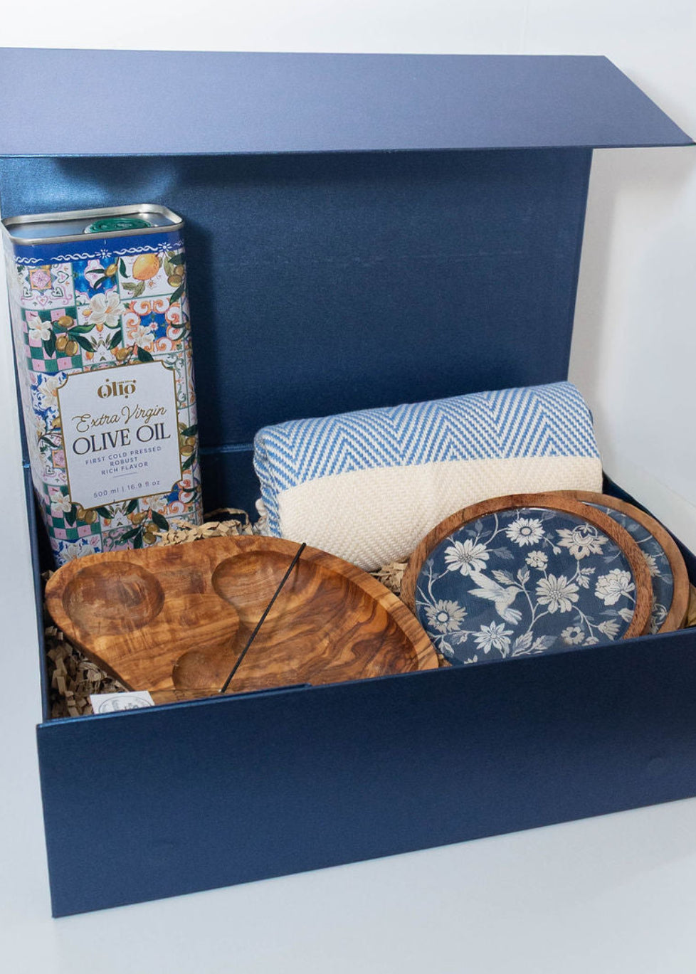 Mediterranean gift box with extra virgin olive oil, two-compartment olive wood serving dish, blue patterned coasters and blue peshtemal towel.