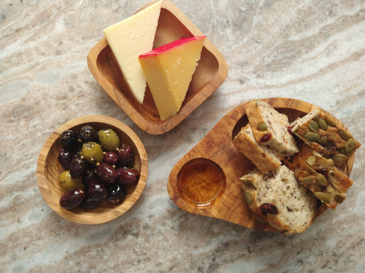 Olive wood serving dishes with assorted cheeses, marinated olives and sliced artisan bread arranged on a marble table.