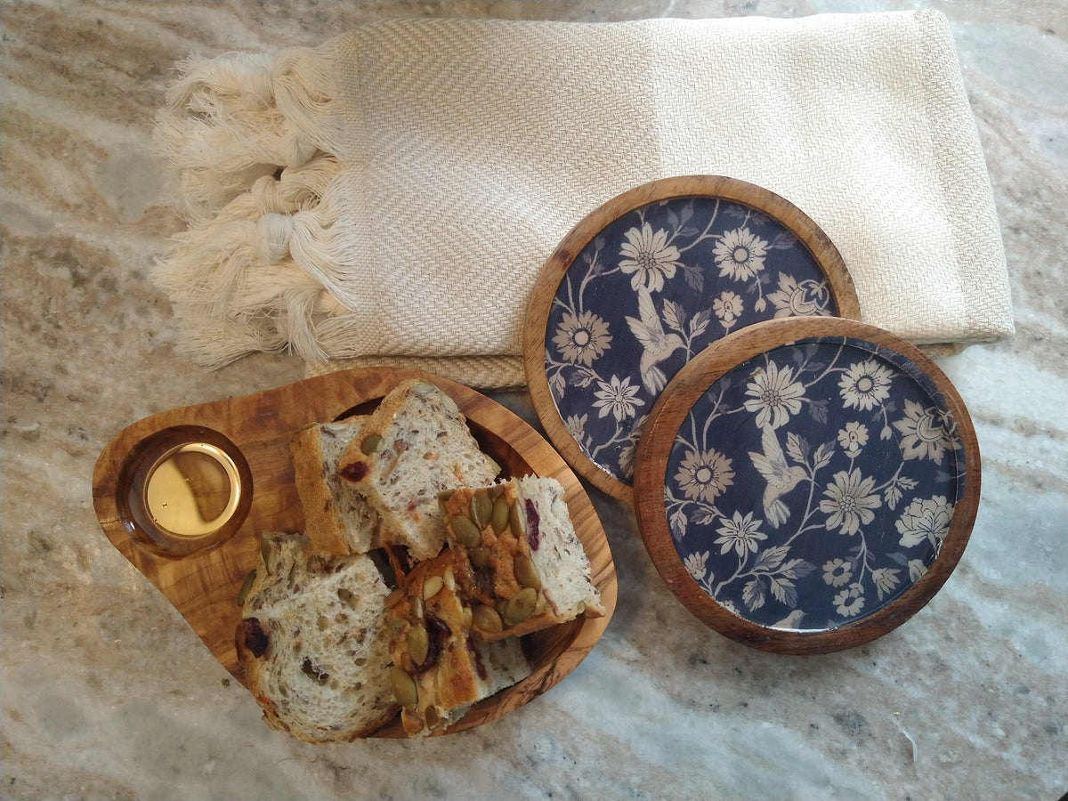 Olive wood serving dish with sliced artisan bread, blue floral coasters and a cream fringed textile arranged on a marble table.