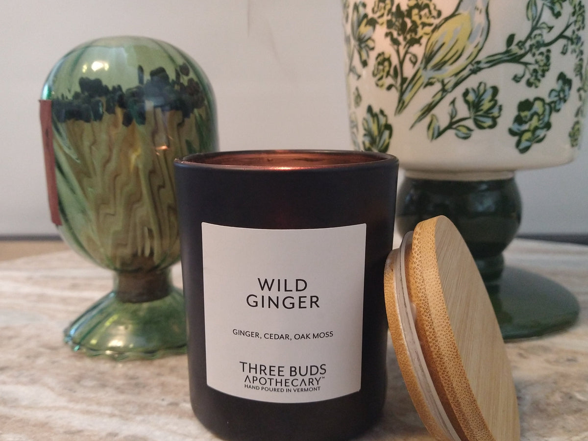 Lit Wild Ginger candle by Three Buds Apothecary beside green glass decor and a botanical table lamp in a cozy home setting.