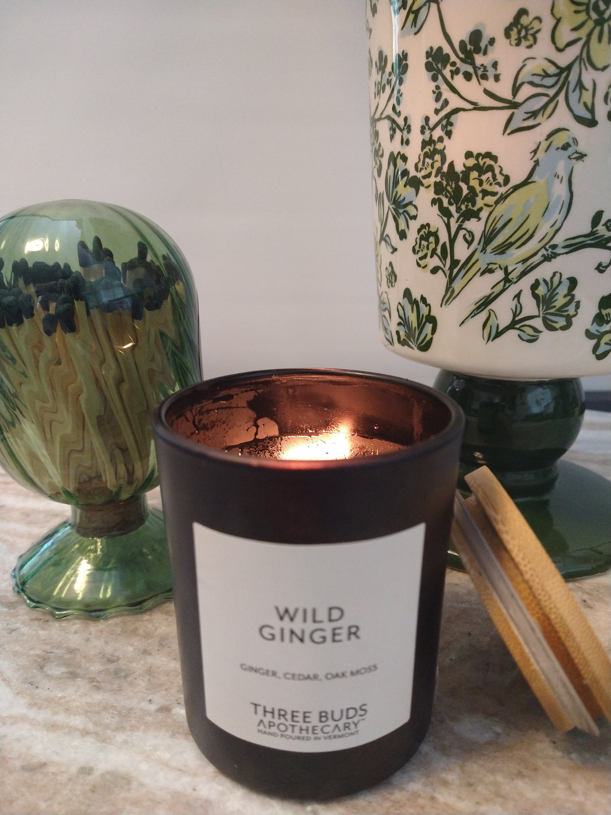 Lit Wild Ginger candle by Three Buds Apothecary beside green glass decor and a botanical table lamp in a cozy home setting.