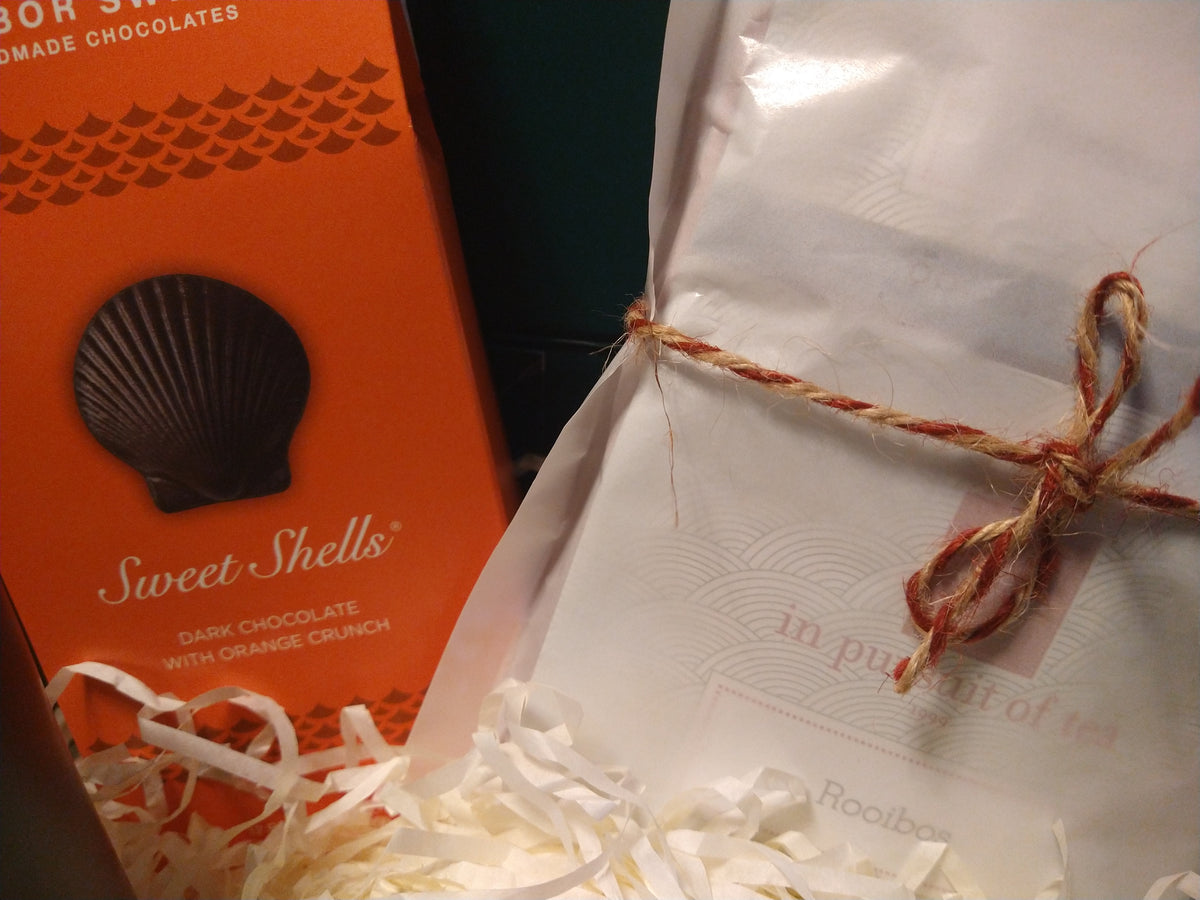 Close-up of the Solefina Autumn Gift Box featuring Harbor Sweets Sweet Shells dark chocolates with orange crunch and an In Pursuit of Tea assortment wrapped in white paper and tied with rustic twine, nestled in cream-colored gift filler.