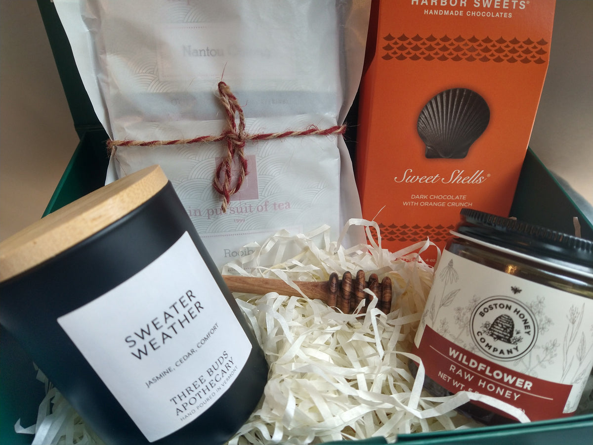 Close-up of the Solefina Autumn Gift Box featuring a Three Buds Sweater Weather candle, Boston Honey Company wildflower honey, an olive wood honey dipper, In Pursuit of Tea assortment tied with twine, and Harbor Sweets Sweet Shells dark chocolates, arranged in a forest-green gift box with cream-colored filler.