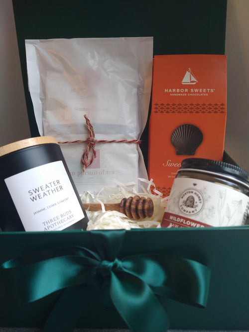Autumn Gift Box in an open forest-green gift box with satin ribbon, featuring a Three Buds Sweater Weather candle, In Pursuit of Tea assortment, Harbor Sweets chocolates, Boston Honey Company wildflower honey, and an olive wood honey dipper.
