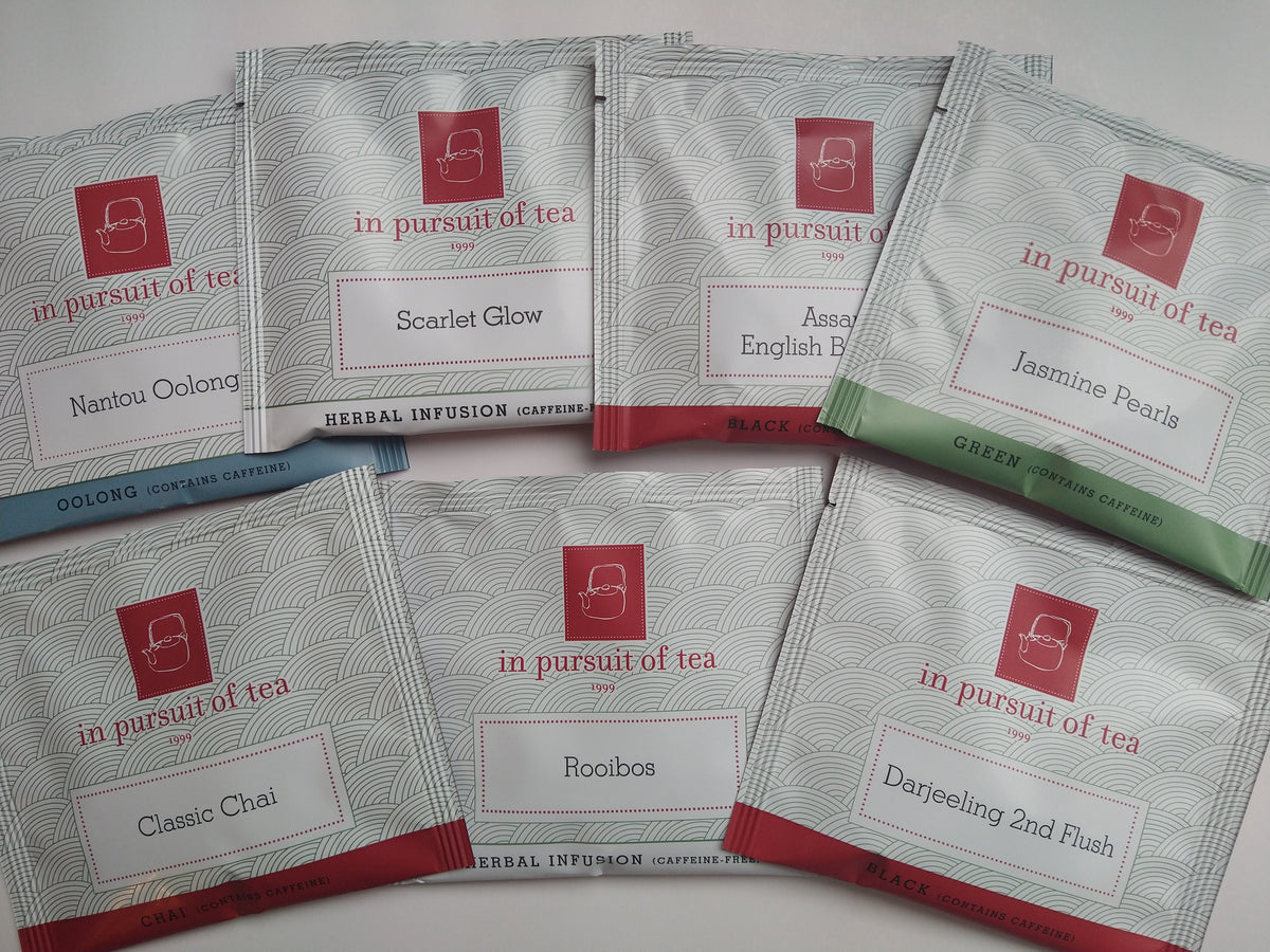 Seven In Pursuit of Tea sachets arranged in a fan, featuring Nantou Oolong, Scarlet Glow, Assam English Breakfast, Jasmine Pearls, Classic Chai, Rooibos, and Darjeeling 2nd Flush teas.