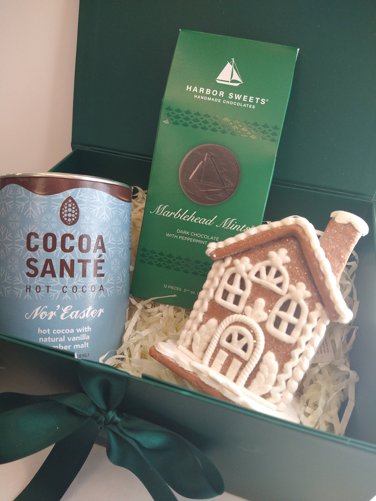 Christmas gift box featuring Cocoa Santé Nor'Easter hot cocoa, Harbor Sweets Marblehead Mints dark chocolates, and a decorative gingerbread house ornament, arranged in an elegant green gift box with ribbon.