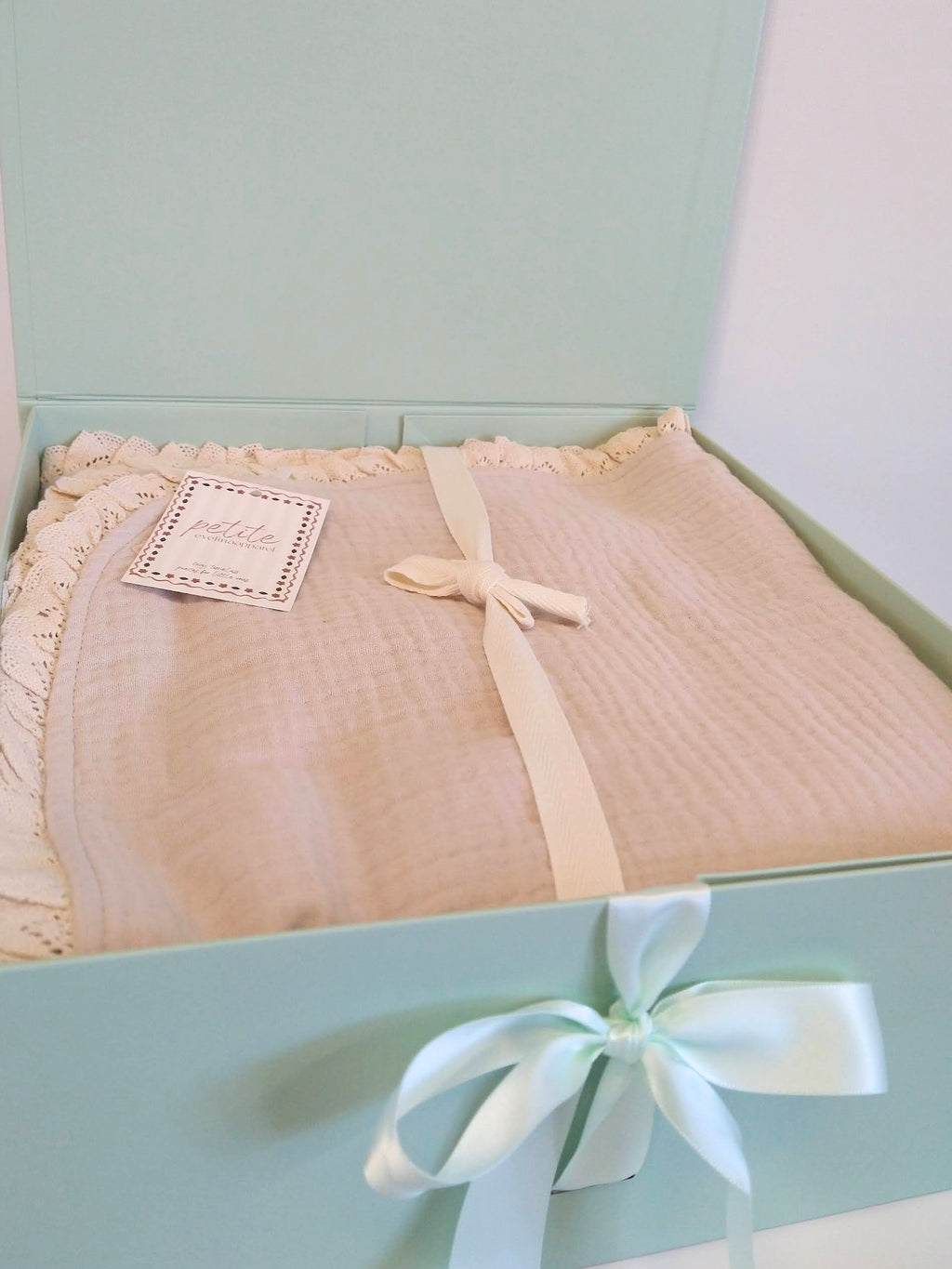 Folded Petite Evelina light beige muslin baby blanket with delicate cotton lace edging and ribbon, presented in an open sage green gift box with satin bow.