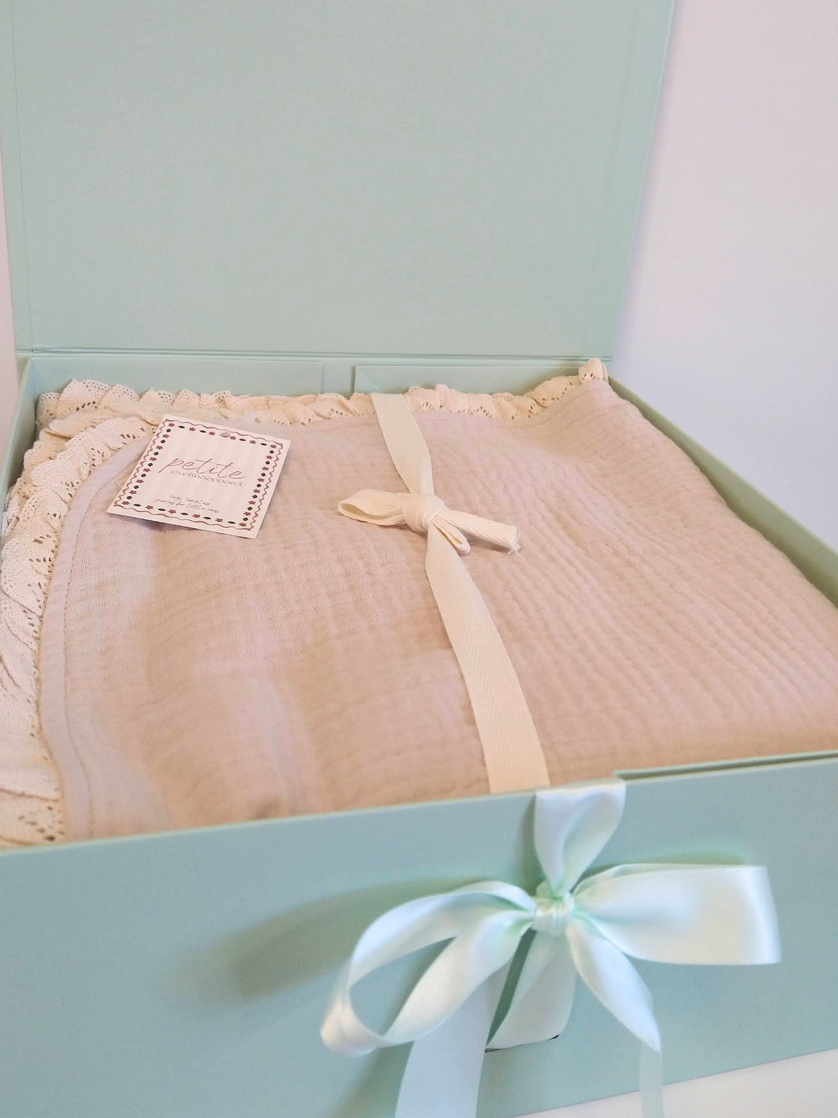 Folded Petite Evelina light beige muslin baby blanket with delicate cotton lace edging and ribbon, presented in an open sage green gift box with satin bow.
