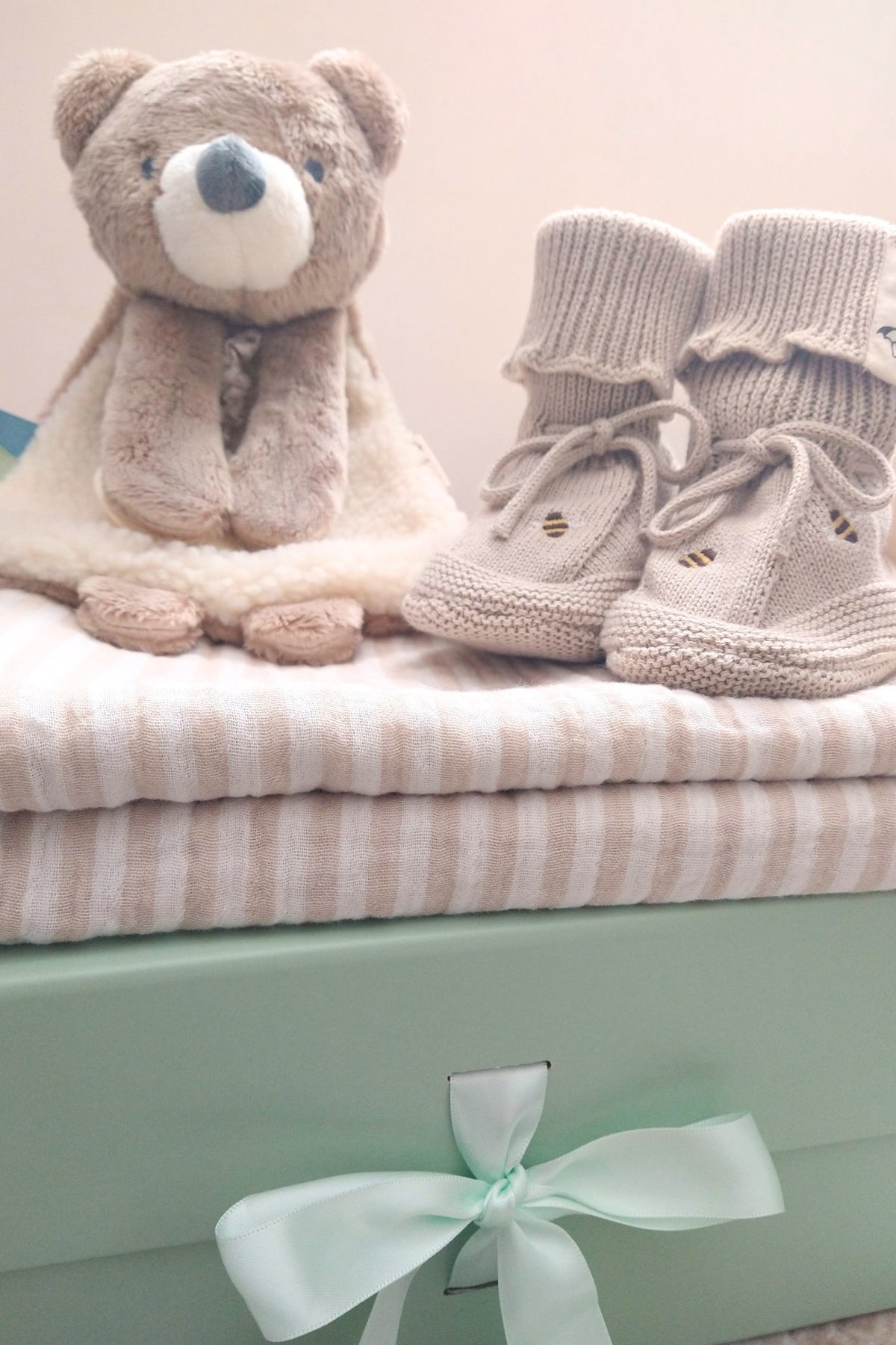 Petite Evelina Beige Stripes baby blanket with Theo Bear Lovey and Saga Copenhagen Honey Bee booties displayed on top of sage green magnetic gift box with coordinated ribbon.