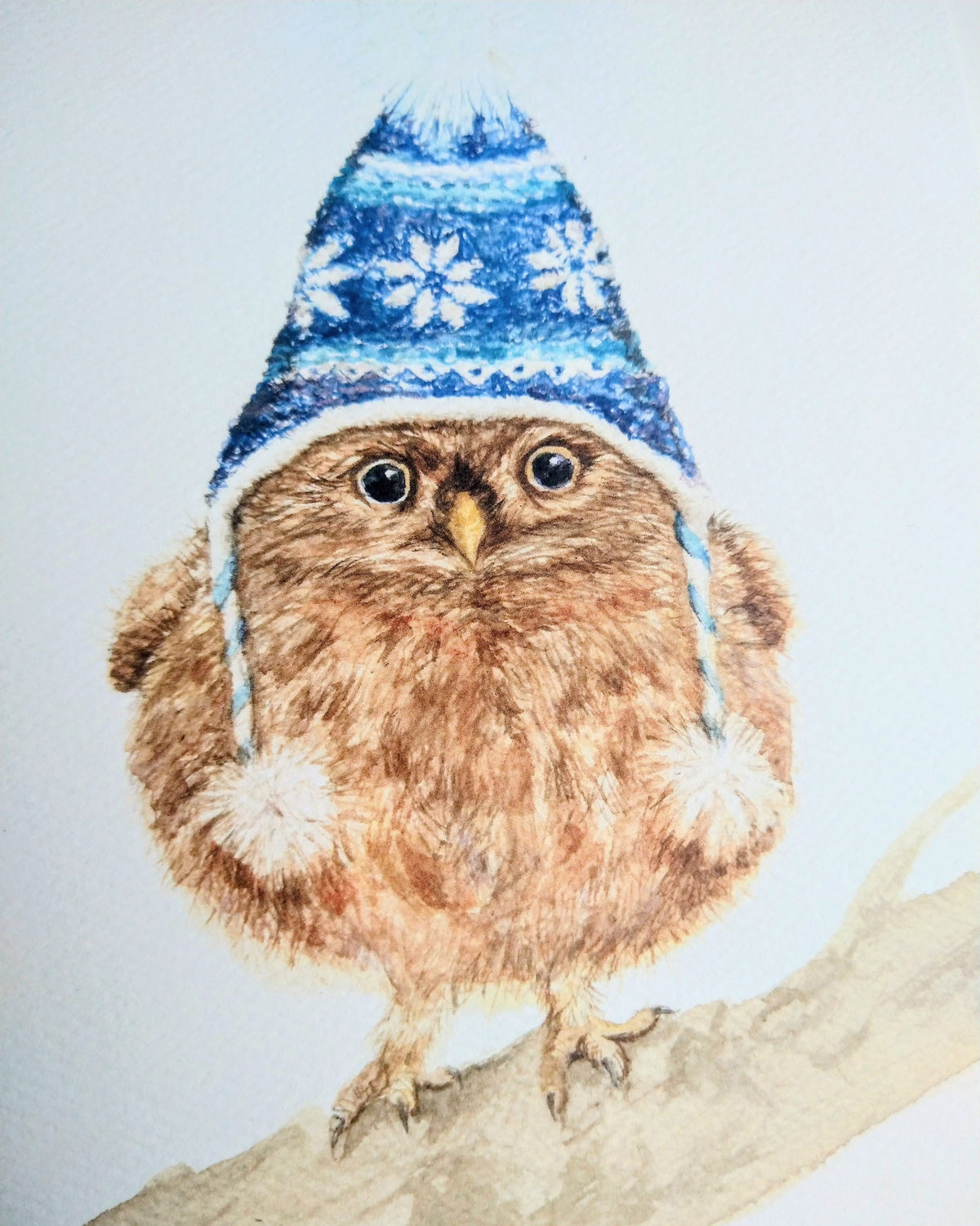 Watercolor illustration of a fluffy brown owl wearing a blue knitted winter hat decorated with white snowflakes and a pom-pom, perched on a branch.